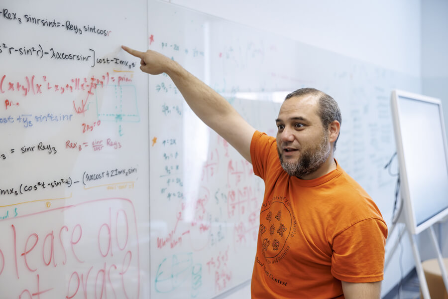 Researcher in orange shirt points to complex mathematical equations written on a whiteboard during lecture or discussion.