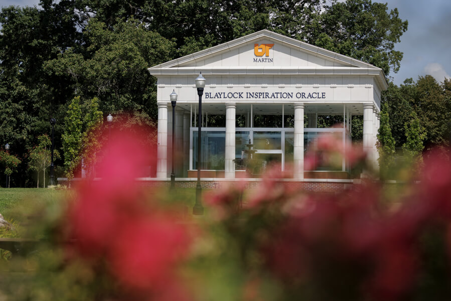 Classical-style white building with columns, "Blaylock Inspiration Oracle" sign, framed by blurry red flowers and trees at UT Martin campus.