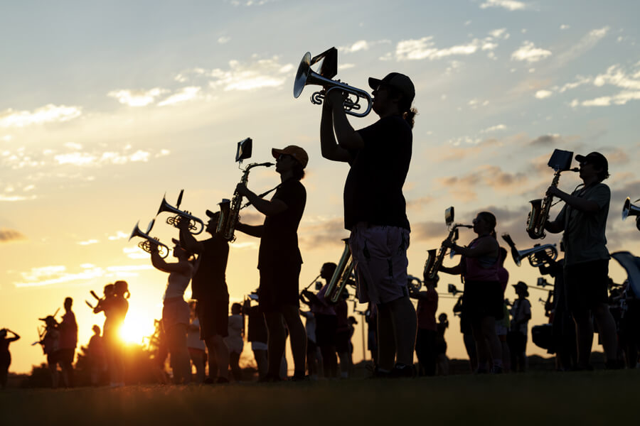 Silhouetted marching band musicians playing brass instruments at sunset, backlit against a golden sky and clouds.