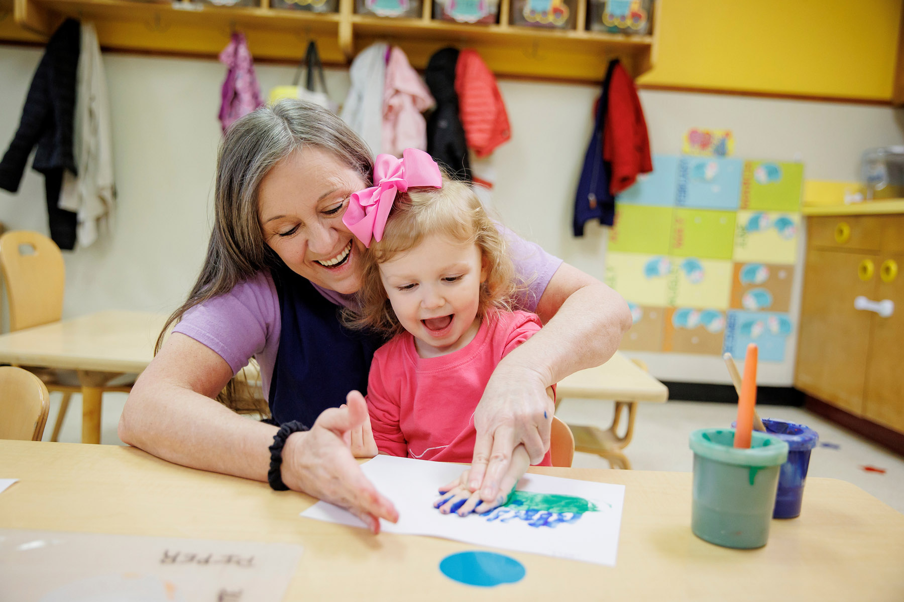 Adult and child joyfully create a colorful handprint painting together in a bright classroom setting with art supplies nearby.